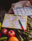 The Faire Meal Planner Notepad, made in the USA, is placed on leafy greens surrounded by apples, oranges, and cucumbers. Two pens rest on top.