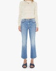 A person stands against a white background wearing The Outside Flood cropped flare vintage blue jeans by Mother, a sheer cream long-sleeve sweater, and dark blue open-toe sandals.