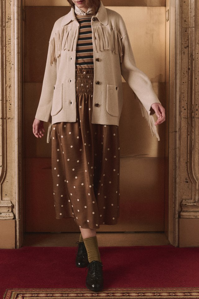 Model in a cream fringed sweater jacket, striped top and copper dotted midi skirt in a wood-paneled doorway.