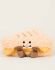 The Faire Grilled Cheese Warmies is a microwavable plush shaped like a smiling grilled cheese, with soft tan "bread," yellow "cheese," brown plush hands and feet, black eyes, and a cheerful smile, providing soothing warmth.