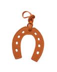 The Faire Bag Charm is a textured orange leather, horseshoe-shaped accessory with square cutouts and a matching hanging loop, adding a stylish, personal touch. Displayed on a white background.