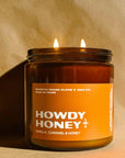 The Howdy Honey Candle by Faire, in a brown glass jar, features vanilla, honey, and caramel notes. Made with coconut soy wax for a long-lasting twin flame burn. Set against tan fabric.