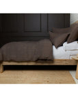A wooden bed topped with the Hunter Duvet and linen shams from POM POM rests against a dark-paneled wall. Nearby, a round wooden table holds books and a vase of flowers on a woven rug.
