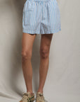 Wearing the Julie Poplin Short by Perfectwhitetee—light blue and white striped cotton with a smocked waist—paired with beige sneakers with green accents, standing against a plain light-colored background for a true high-summer staple look.