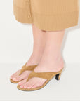 A person in a tan skirt stands against a white background, showing only their legs and feet. They wear Freda Salvador's KAT—tan suede heeled thong sandals with open toes, evoking classic '90s Italian leather style.