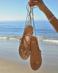 A hand holds the Beek Koha Sandal, featuring thin brown leather ankle-tie straps, dangling in front of a serene beach with gentle waves and a clear blue sky.