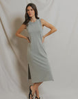 A woman with dark wavy hair stands before a light fabric backdrop wearing the Perfectwhitetee Lanell Cotton Tank Dress in light gray, a sleeveless midi with side slits, paired with black flip-flops and her arms on her hips.