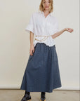 A woman poses against a plain background in a loose white button-up shirt, Aquarius Cocktail Le Cargo Skirt in blue plaid Japanese cotton poplin, argyle socks, gold pointed heels, and a chain belt at her waist.