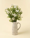 A white ceramic pitcher holds green foliage and Indaba's 01-9528 Lotus Flower Branches, creating elegant accent decor against a plain beige background.