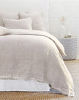 In a cozy bedroom scene, a neatly made bed is adorned with beige linen bedding and matching pillows from the Logan Duvet by POM POM. A textured rug covers the floor, and a round side table displays a vase and candle, all set against a white brick wall for that perfect heathered effect.