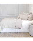The minimalist bedroom features a neatly made bed adorned with POM POM's Logan Duvet in light beige linen, complemented by pillows and a textured bedspread. Next to the bed is a round, woven side table set against walls with subtle panel detailing. The floor showcases a natural woven rug from the Loga Collection, offering a heathered effect.