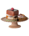 The Maileg Cakes Display by Maileg features a fluted pink pedestal cake stand holding three assorted pastries: a square chocolate petit four with red berry and two round mini tarts with cream and purple garnish.