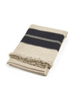 The Marshall Throw by Libeco Home is a folded beige wool blanket featuring a wide black stripe and flax fringe on one edge, displayed against a white background.