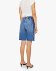 A woman stands with her back to the camera, wearing a white short-sleeve shirt and Mother’s Mid Rise Maven Bermuda Fray shorts with a frayed hem, paired with black heeled sandals against a plain background.