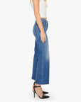 A woman in profile wears Mother’s The Mid Rise Maven Flood—mid-rise, wide-leg blue stretch denim jeans—paired with a white sleeveless top and black heels against a plain white background.