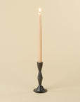 A lit beige taper candle stands in the Indaba 1-2506 Zora Forged Candlestick L, featuring a dark metal finish, set against a light beige background.
