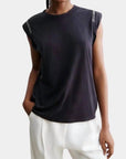 A person wearing the Rachel Comey Miles Tee with white text on the shoulders and loose-fitting white pants stands with one hand in a pocket against a plain background.