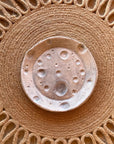 Faire’s Moon Trinkets rest in a ceramic dish featuring a speckled, cratered surface that resembles the moon, displayed on a fringed woven placemat—perfect for showcasing your favorite celestial décor.