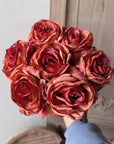 A close-up of a hand holding the Faire 17'' Faux Vintage Rose Bouquet, featuring red and orange blooms with dried, curled petals, set against a blurred wooden background.