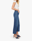 A woman stands in profile against a plain white background, wearing The Half-Pipe Ankle high-waisted jeans in a dark blue wash by Mother, paired with a short-sleeve white top and navy heeled sandals.