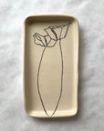 The Floral Small Tray by Faire is a rectangular cream ceramic tray featuring a minimalist black line drawing of two poppy flowers, one large and one small, etched by hand. It is displayed on a white textured surface.