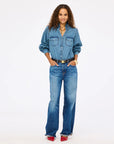 A woman stands confidently in a Kerri Rosenthal Puff Sleeve Shirt tucked into high-waisted blue jeans, hands in pockets. She finishes the look with a black belt, red shoes, layered necklaces, and a gold watch, smiling against a plain background.