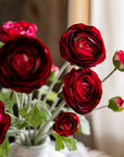 The Buttercup Flower Bundle Red Burgundy by Faire features deep red faux silk flowers with green leaves in a white vase, gently illuminated by natural window light.