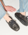 A person wearing light blue jeans and Freda Salvador RIA Driver Loafers in black Nappa leather with thick soles sits on a light surface, legs extended, hands resting on the floor.