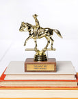 A gold-colored trophy of a cowboy on a horse, mounted on a wooden base with a plaque that reads "THIS AIN'T MY FIRST RODEO," is placed on top of a stack of books with orange and white covers. Perfect for adults in Winston Salem who love to win stuff, the JE Trophy by Faire is an ideal prize.