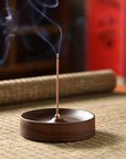 The Faire Walnut Wood Incense Burner emits a thin trail of smoke, placed beside handcrafted ceramic plates featuring traditional Chinese designs. The setup sits on a woven mat with blurred bamboo and red panel backgrounds.