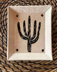 The Saguaro Soap Dish is a handmade sgraffito piece with a black cactus design at its center and four drainage holes, presented on a woven circular mat featuring brown and black patterns.