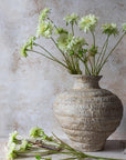 A textured beige ceramic vase holds Abigail Ahern's Scabious Flower, Cream—long-stemmed blooms with green centers. A single stem and a few cut flowers, plus scissors, rest on the table against a light stone background.