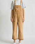 A model showcases the Denimist RELAXED OVERALL—a tan, 100% cotton piece featuring adjustable buckle straps—layered over a loose gray T-shirt and paired with white sneakers. Against a plain white background, the overall’s relaxed-leg silhouette highlights its laid-back style as the model stands with arms comfortably at their sides and face partially out of frame.