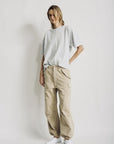 A person stands against a plain white background wearing a relaxed-fit light gray t-shirt, Denimist CROPPED CARGO beige utility pocket pants, and white shoes, with one hand in their pocket.