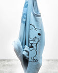 A BasicGoodness Schulz Raglan by Free City (sparrow, LLC), in light blue and made in the USA, features a black outline graphic of Snoopy hugging Woodstock. It hangs on a plain, light background.
