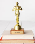 A JE Trophy styled like a human figure stands atop a stack of books with a plaque reading "STAY GOLDEN" on a white background.