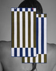 A grayscale photo of a person’s bare back and shoulders with two Typical Navy/Moss Stripe Bath Towels digitally superimposed in blue, olive green, and white over their upper back and hair.