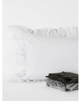 The Tat Cotton Sham Euro by Matteo, in white cotton with ruffled edges, is displayed against a white background. In front, gray and black fabric items highlight the soft percale weave and 225 thread count.
