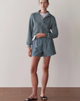 A person stands indoors against a plain light wall and wooden floor, wearing Donni's The Stripe Pop Boxer in blue-and-white stripes, paired with a light blue pullover, white t-shirt, and dark slide sandals.