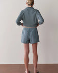 A person stands facing a blank wall, wearing a cropped, long-sleeve gray top, Donni's The Stripe Pop Boxer in blue and white stripes, and black sandals. Their hair is pulled back in a low bun.