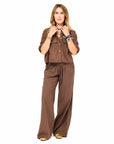 Against a white background, a woman with blonde hair wears the Kerri Rosenthal Tia Love Brigade Shirt and matching wide-leg pants. She pairs them with gold shoes and accessorizes with bracelets and necklaces.