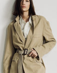 A woman with straight brown hair wears the Denimist DECONSTRUCTED BLAZER WITH BELT in beige over a white pinstriped shirt, standing against a plain light background and looking at the camera with a neutral expression.