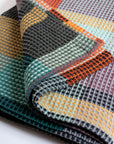 A close-up of folded Vrou-Vrou fabric by Faire highlights its bold waffle texture and colorful geometric pattern in orange, yellow, teal, black, and purple.
