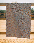The Winchester Orange Quilt by Faire, ideal for cozy quilts, showcases three sections of colorful floral patterns and is displayed draped over a wooden fence outdoors with sandy ground and blurred trees in the background.