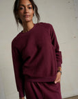 A woman stands indoors wearing the Perfectwhitetee Ziggy Reverse LS Sweatshirt and matching pants in textured maroon cotton fleece, gazing calmly at the camera against a plain light background.