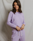 A woman with long brown hair stands in front of a light backdrop, wearing the Perfectwhitetee Ziggy Reverse LS Sweatshirt in light purple with a shrunken fit and bracelet-length sleeves, paired with matching drawstring sweatpants.