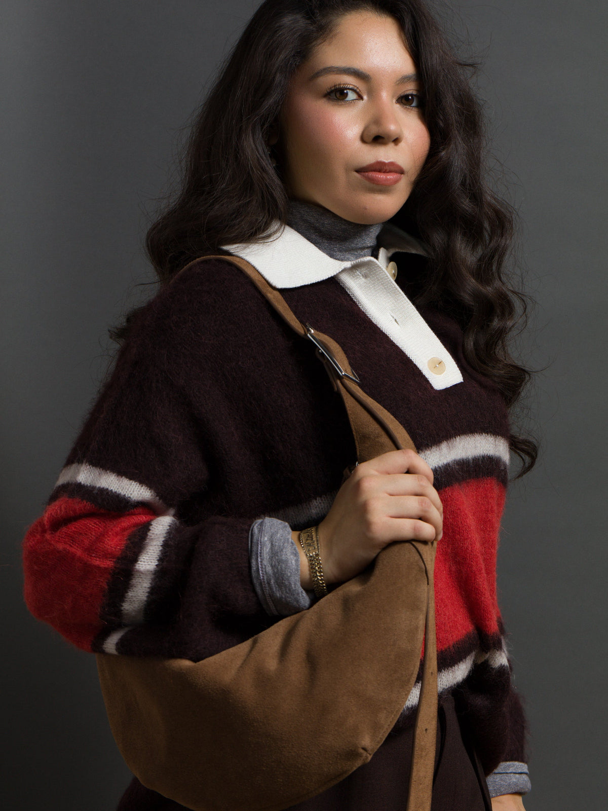 A person with long dark hair wears a striped polo sweater over a gray turtleneck and carries a brown shoulder bag.