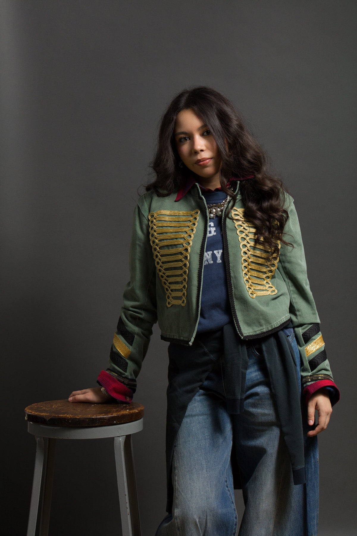 A young woman with long wavy hair wears a cropped olive-green military-style jacket with gold embroidery over a blue graphic t-shirt and jeans, posing with one hand resting on a wooden stool.