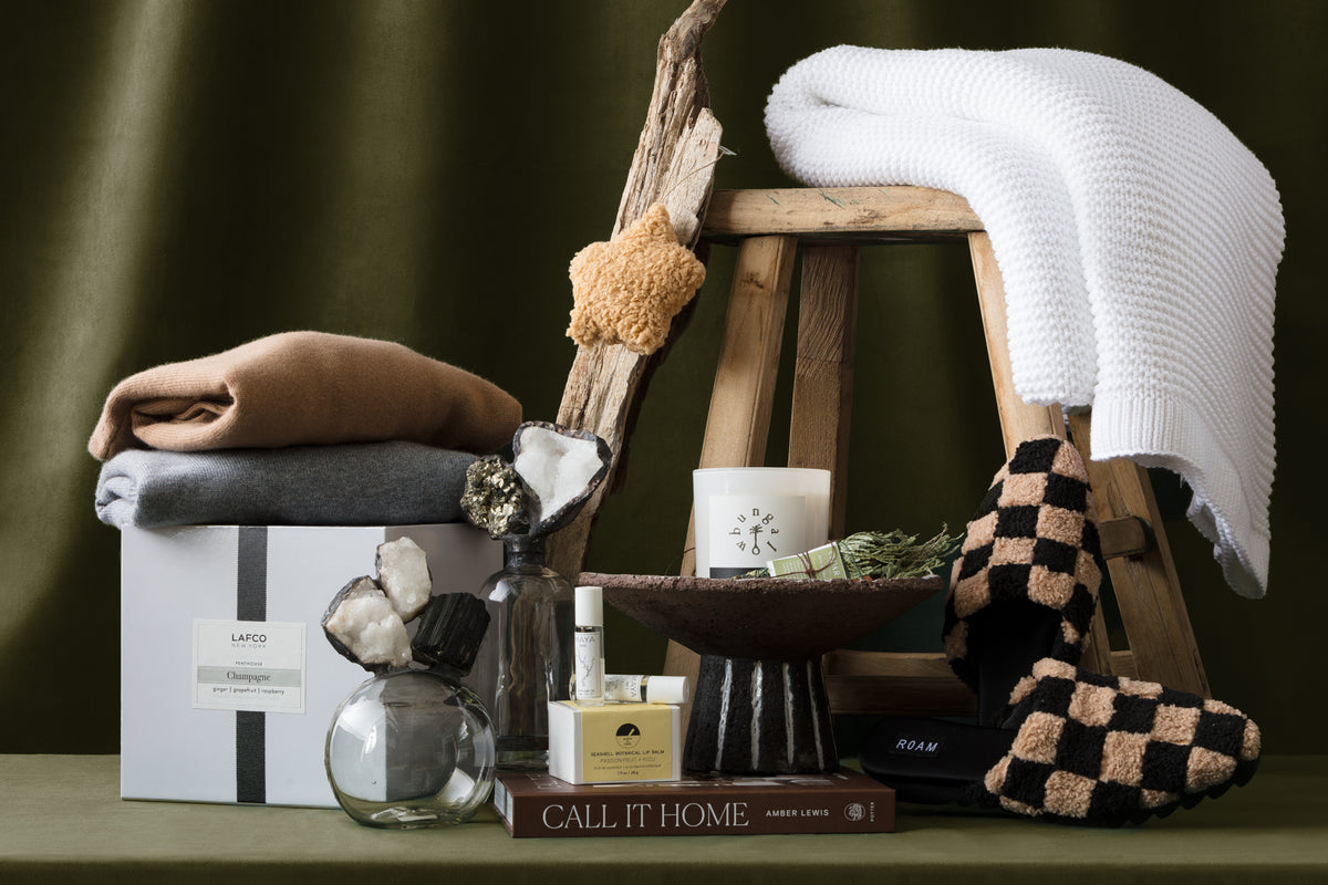 A cozy display featuring folded sweaters, a white knit blanket, slippers, a scented candle, books, skincare products, crystals, and a vase arranged on a wooden stool against an olive green background.
