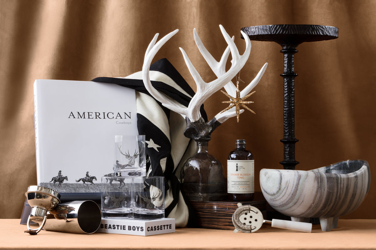 A stylish arrangement features a book titled "American Cowboys," striped cloth, white antlers, a star ornament, candle, marble bowl, cocktail items, glassware, a bottle, and a Beastie Boys cassette.